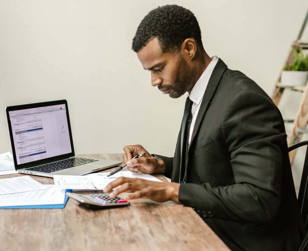 Black businessman in a suit working at a desk with a laptop, calculator, and papers in a modern office